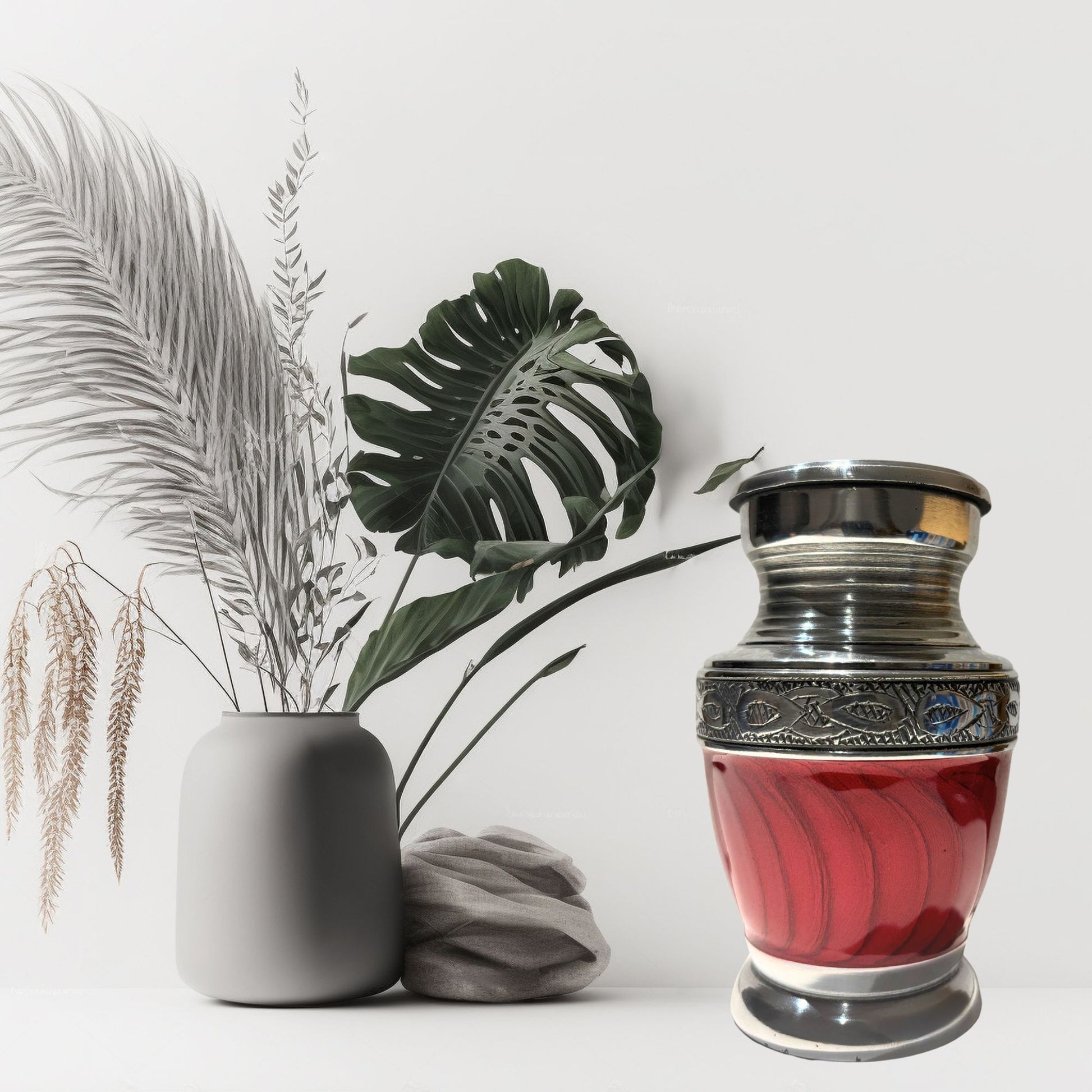 Silver urn with red interior next to a gray vase and decorative leaves on a white background
