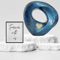 Blue marble-like sculpture with a heart detail on a white pedestal, accompanied by a framed quote 'Forever with us' on a light gray background.