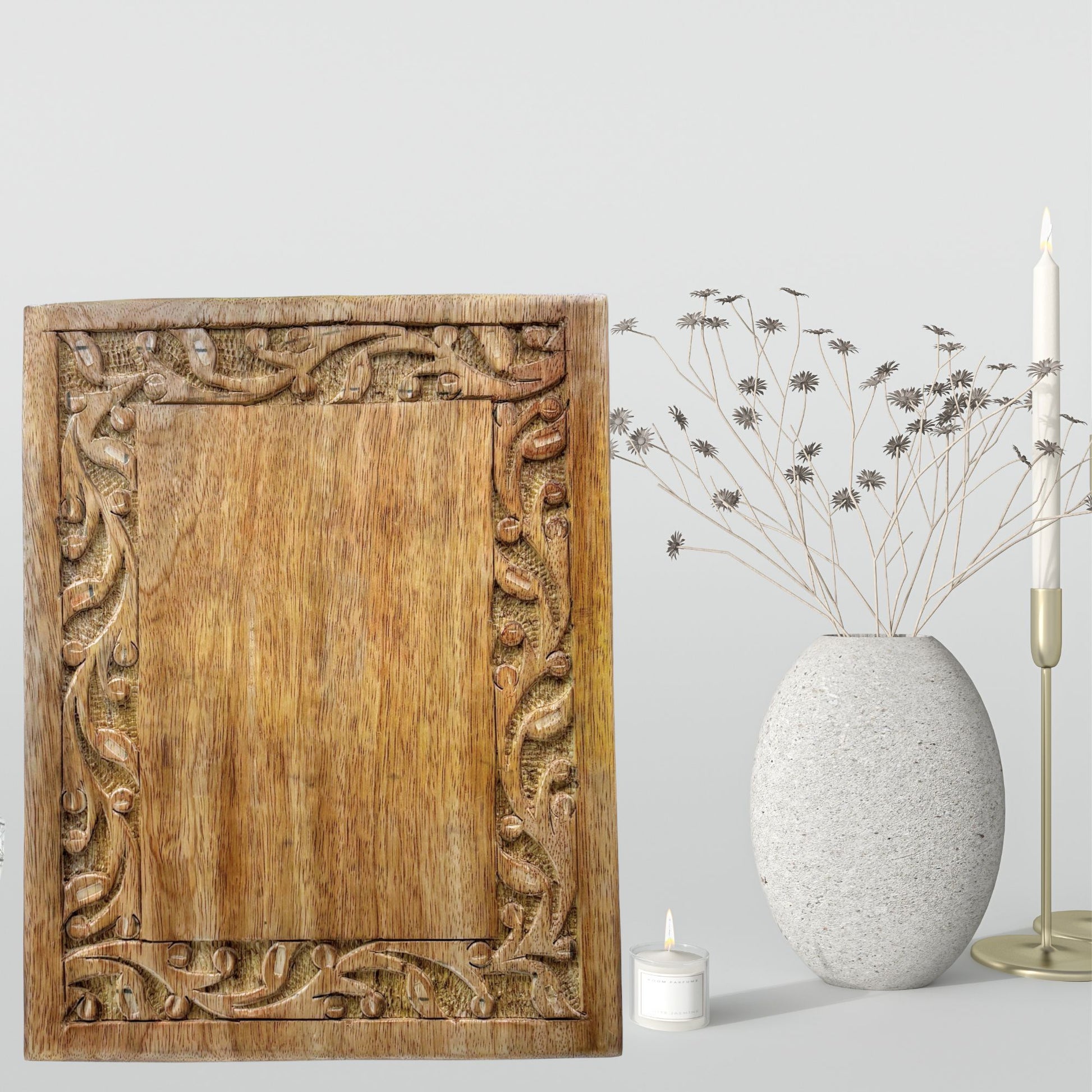 Carved wooden panel with decorative patterns next to a vase with dried flowers and a candle on a light background