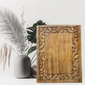 Wooden decorative panel with intricate carvings next to a vase and plants on a white background
